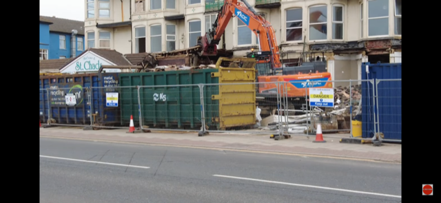 A building being demolished with a green skip from Kirkby Skips in front and behind guard rails. Between the building and the skip is crane.